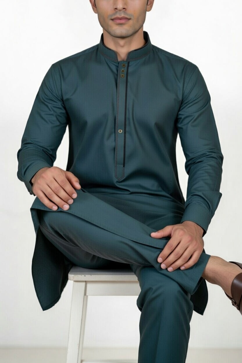 Man wearing a teal traditional outfit sitting on a white stool against a white background