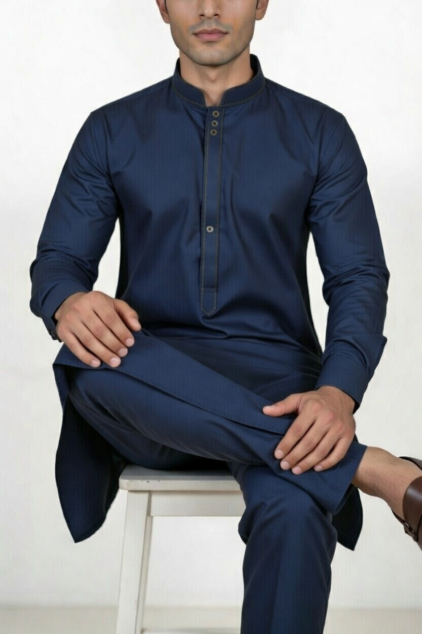 Man wearing a navy blue traditional outfit sitting on a white stool against a white background