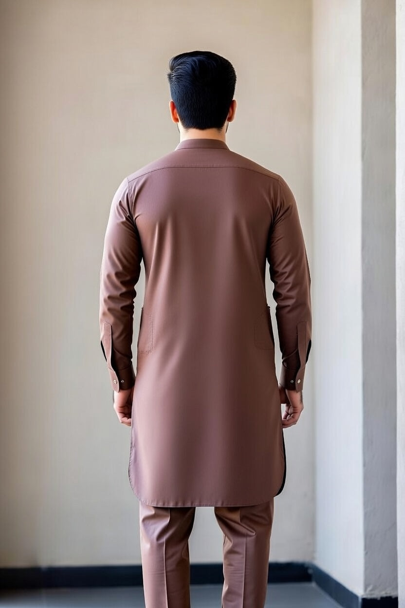 Man wearing a brown traditional outfit standing against a plain wall.