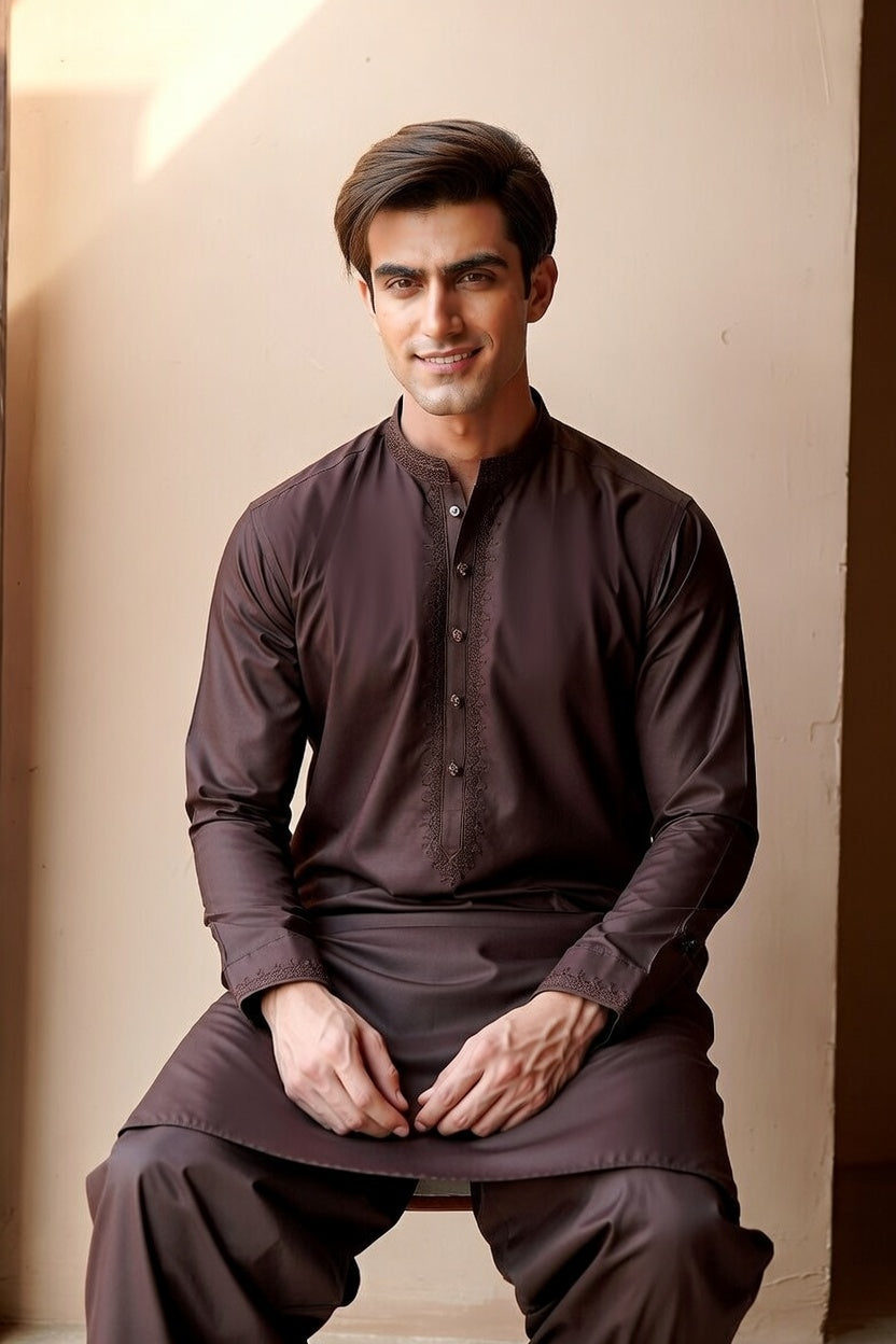 Man wearing a dark brown traditional outfit sitting against a beige wall.